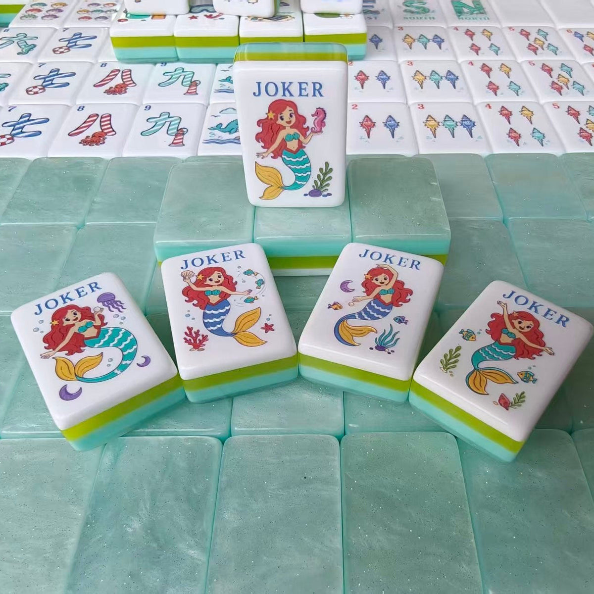 Scattered pile of Mermaid Ocean mahjong tiles, showing mermaid joker, whale, conch shell, seahorse, West wind tile and beach hut artwork, mint green translucent backs with green side stripes