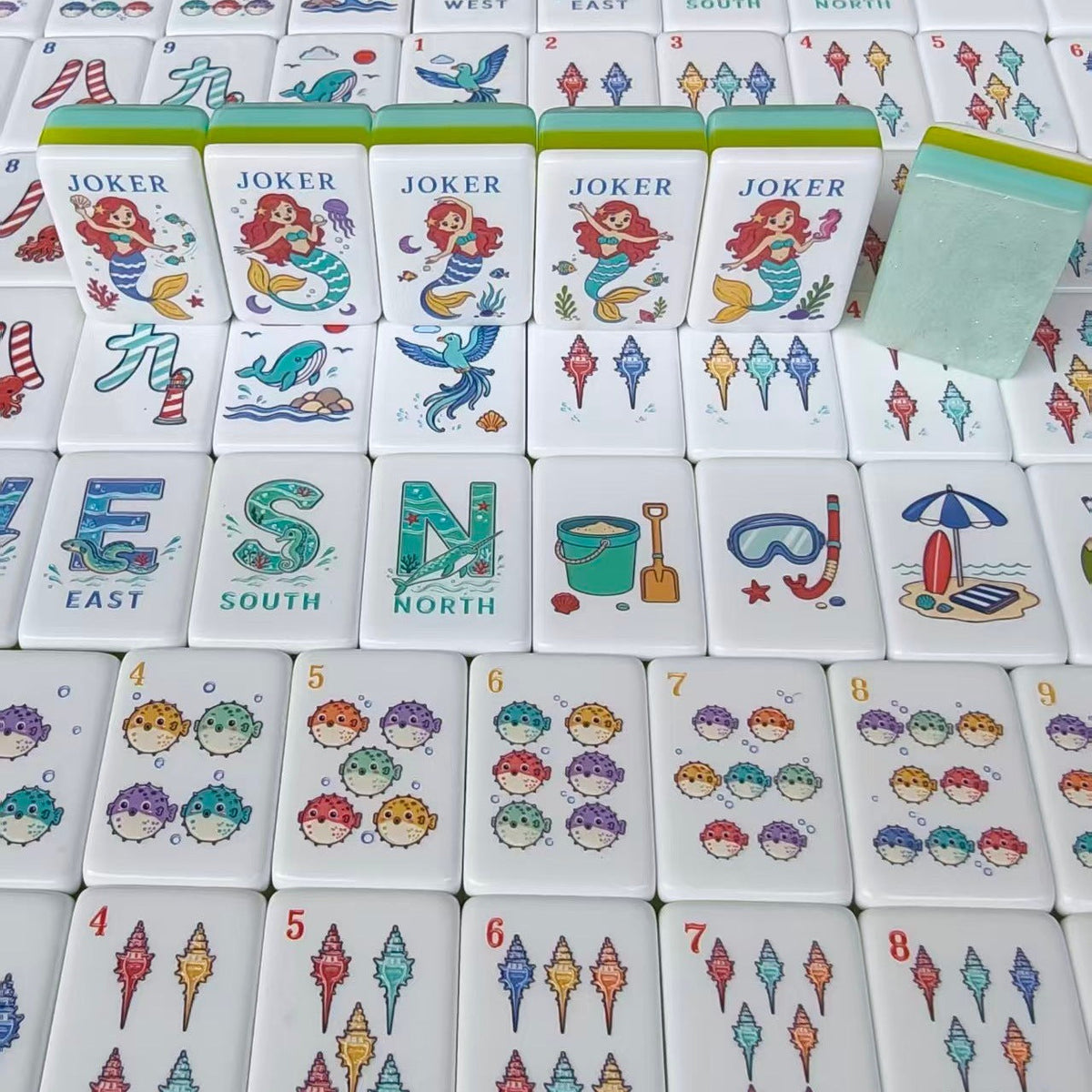 Overhead scattered shot of Mermaid Ocean mahjong tiles, showing South wind tile with seahorse, West wind with whale, North wind, ship wheel crack tiles, seahorse crack tiles, mermaid jokers and beach hut tile