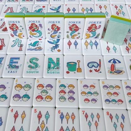 Overhead scattered shot of Mermaid Ocean mahjong tiles, showing South wind tile with seahorse, West wind with whale, North wind, ship wheel crack tiles, seahorse crack tiles, mermaid jokers and beach hut tile