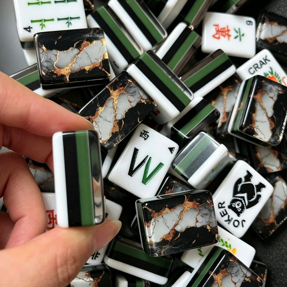 Hand holding mahjong tile displaying black marble back with white stone veins and shimmer gold foil, pile of black gold marble mahjong tiles in background