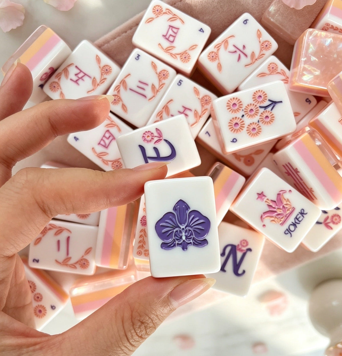 Hand holding Orchid Crown Pink mahjong tile with purple orchid design, scattered mahjong tiles with rose gold floral art, blush pearlescent backs, soft pink fabric background