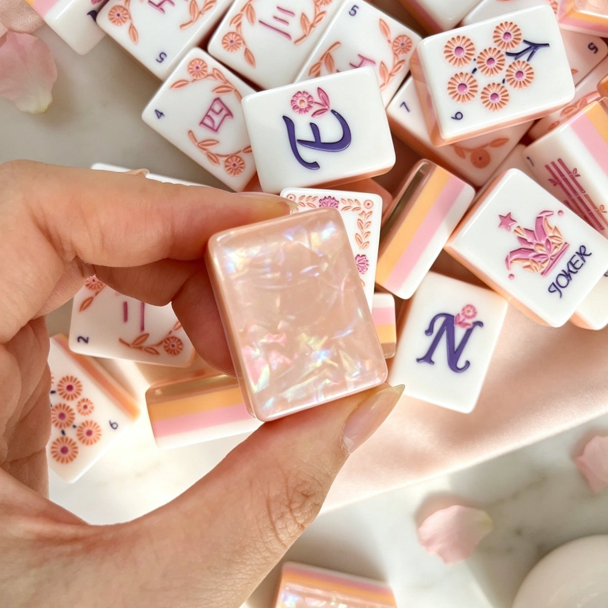 Hand holding Orchid Crown Pink mahjong tile with purple orchid design, scattered mahjong tiles with rose gold floral art, blush pearlescent backs, soft pink fabric background