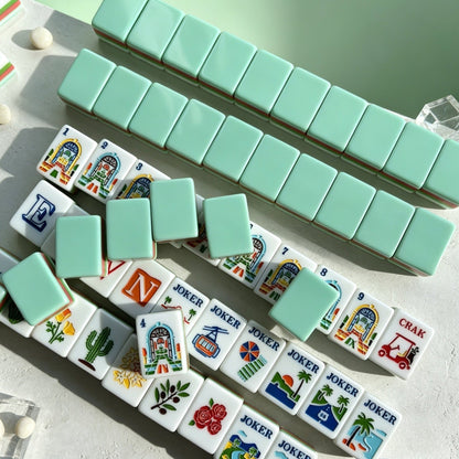 Overhead shot of scattered tropical golf resort mahjong tiles, mint green backs, pink and green striped edges, vacation themed artwork on white tile faces, styled on light green background
