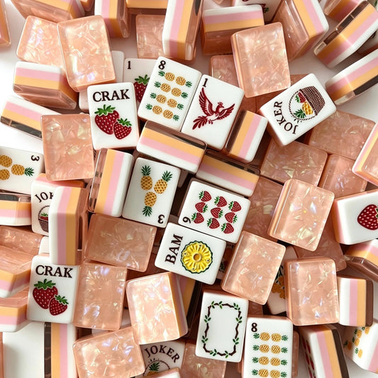Tropical Fruit Garden American mahjong tiles scattered on marble table, pink shimmer crushed shell backs, pastel layered side stripes, strawberry and pineapple fruit illustrations, cream silk fabric backdrop