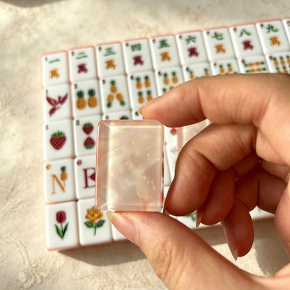 Scattered pile of Fruit Garden mahjong tiles, shimmering pink pearlescent backs, pastel pink-orange layered edges, strawberry and pineapple illustrated tile faces on linen surface