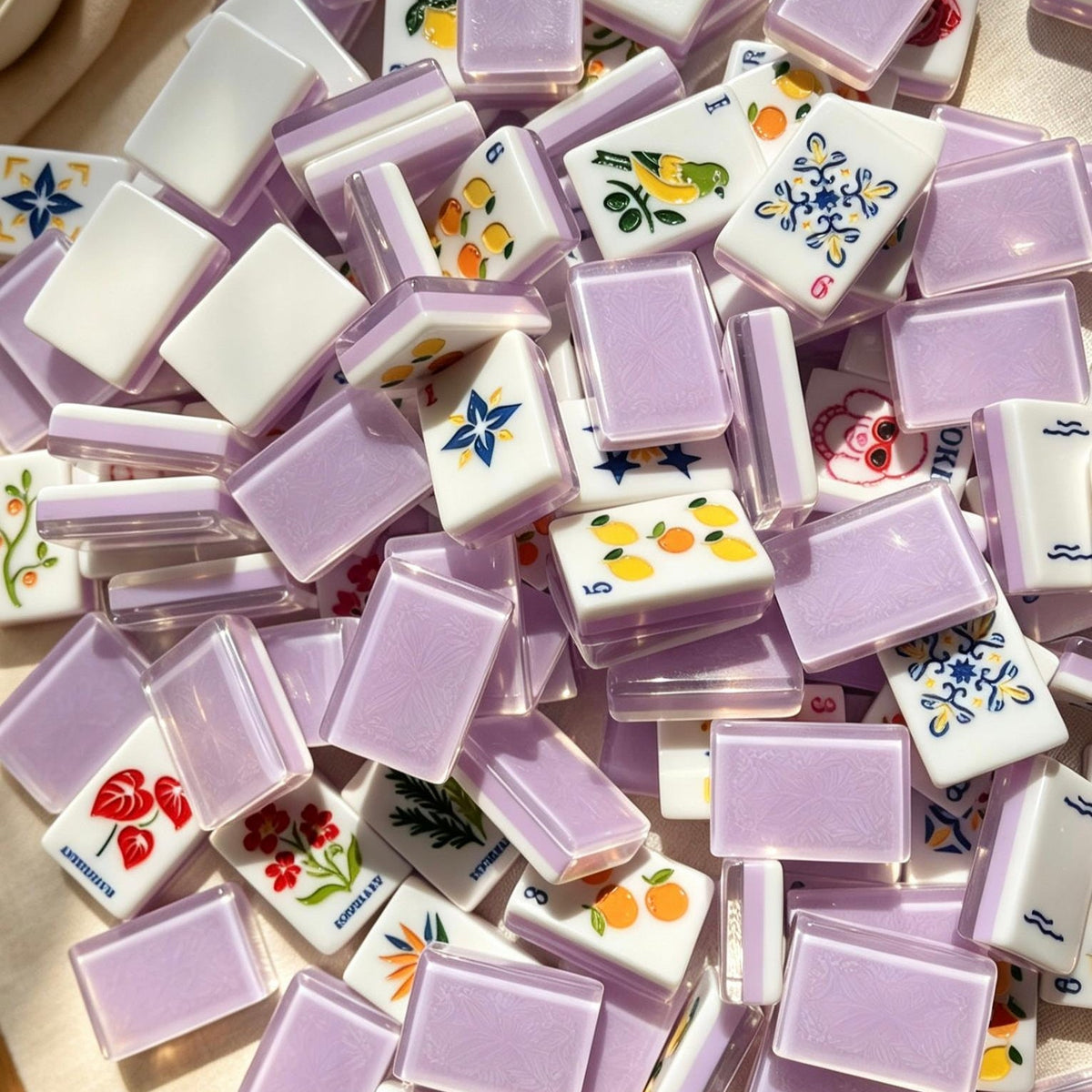 Lavender Fruit & Floral American mahjong tiles, translucent lavender purple resin backs, illustrated citrus fruit, floral and cartoon joker tiles, arranged on cream marble countertop with minimalist vases