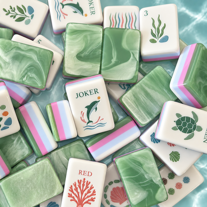 Top-down flat lay of full Coastal Breeze mahjong set laid out on water ripple background, white tile faces with ocean illustrations: seahorse, seashell, coral, dolphin joker, turtle dragon, lighthouse wind tiles, one green marbled tile at corner