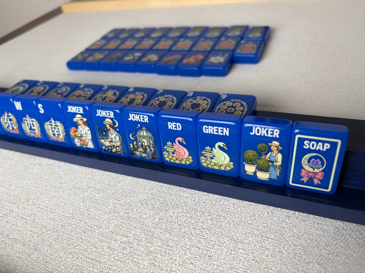 Side profile shot of Blue Chinoiserie mahjong tiles lined on dark wooden rack, glossy cobalt tiles displaying garden joker scenes, pink and white swan dragon tiles and floral SOAP tile